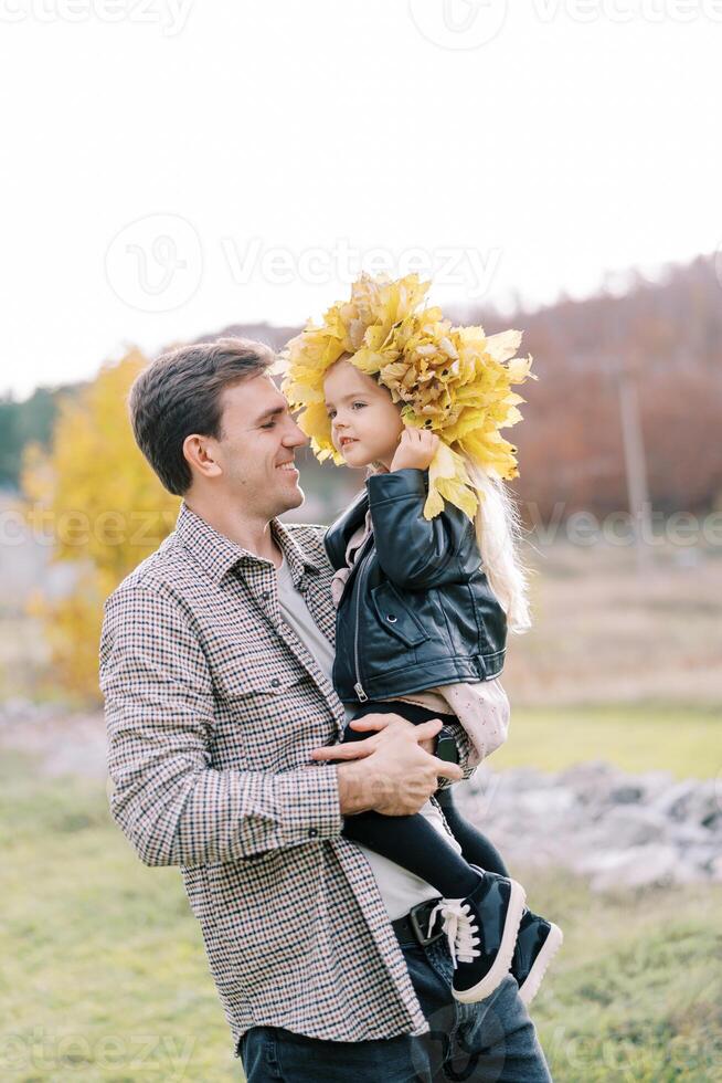 papà con un' poco ragazza nel un' ghirlanda di giallo le foglie nel il suo braccia sta nel un' radura foto