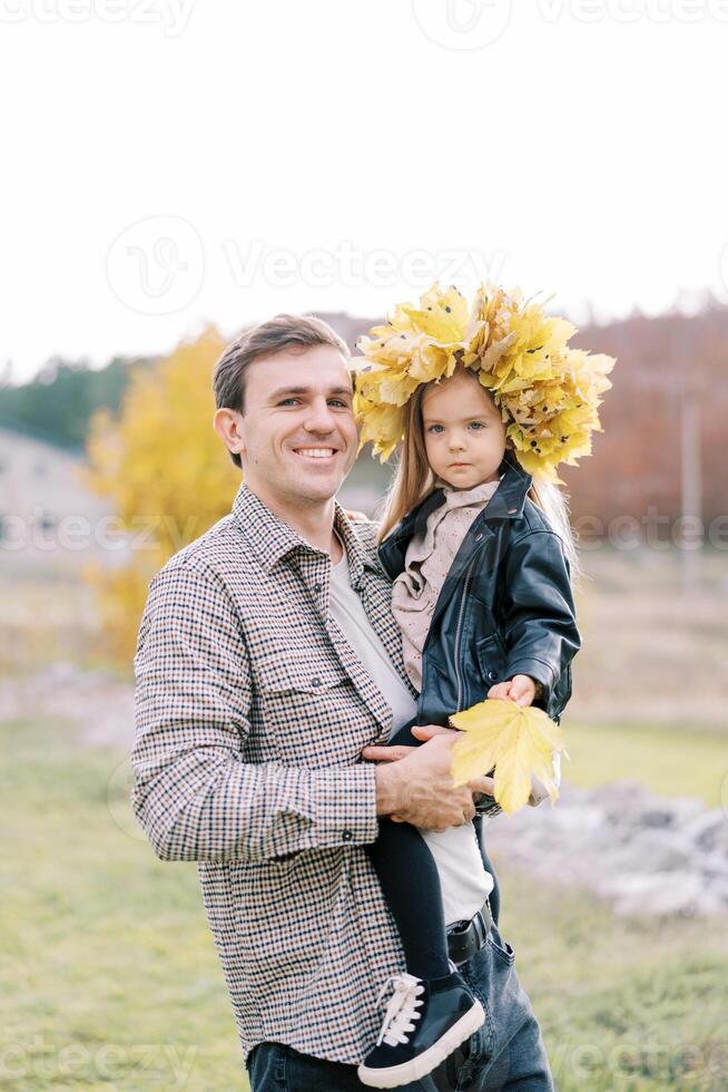 sorridente papà Tenere nel il suo braccia un' poco ragazza nel un' ghirlanda di autunno le foglie con un' giallo foglia nel sua mano foto