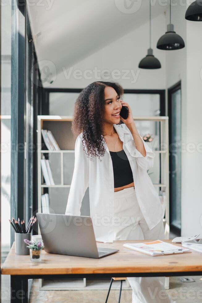 donna d'affari nel un' rilassato posizione chat su il Telefono, sua espressione uno di piacere e Fidanzamento, nel sua moderno ufficio con un' il computer portatile Aperto prima suo. foto