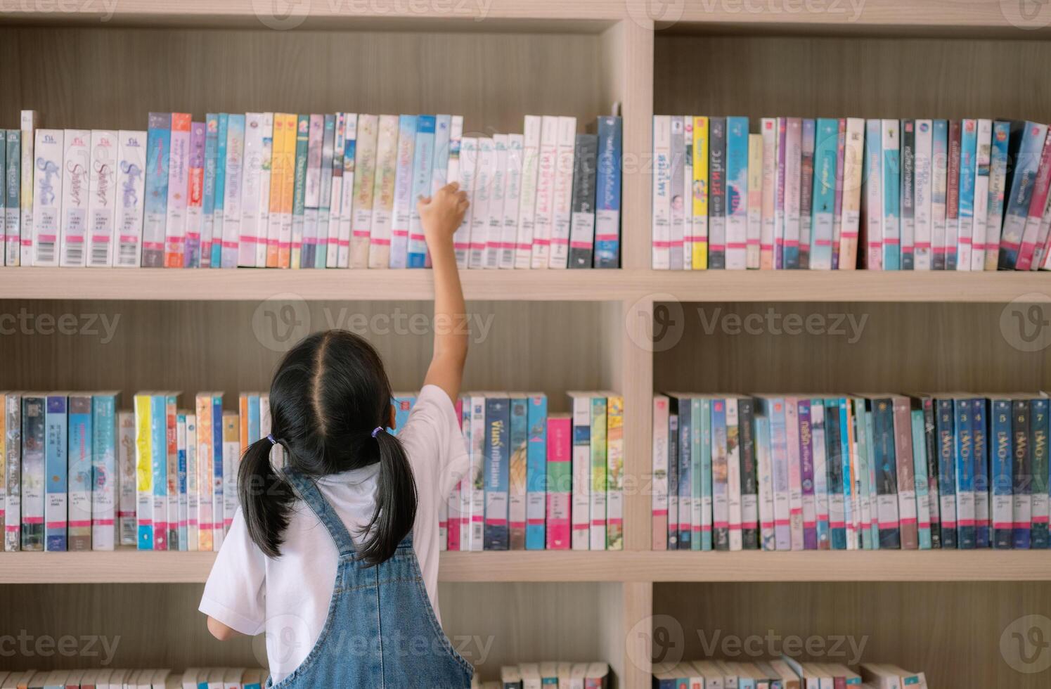 un' giovane ragazza è raggiungendo per un' libro su un' biblioteca mensola foto