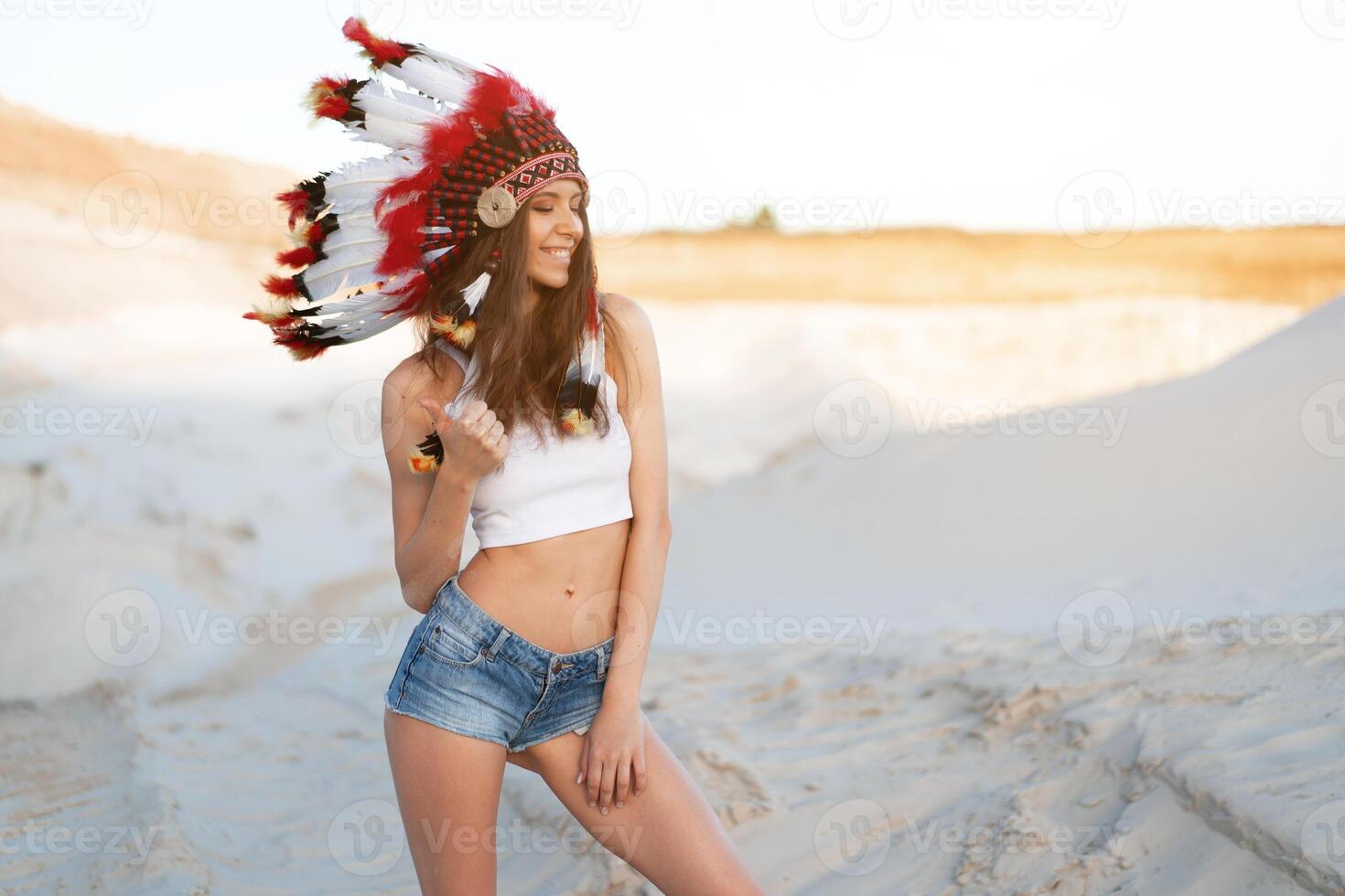 un' bellissimo giovane caucasico ragazza nel un' bianca superiore e denim corti. su sua testa indossare un indiano cappello scarafaggio. in piedi nel il deserto. foto