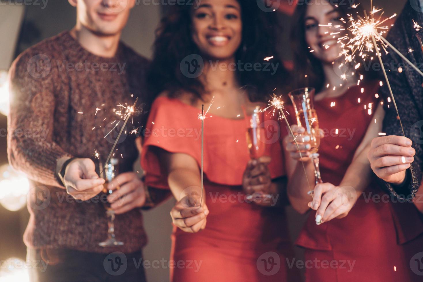 foto a fuoco. amici multirazziali festeggiano il nuovo anno e tengono in mano luci e bicchieri del Bengala con un drink