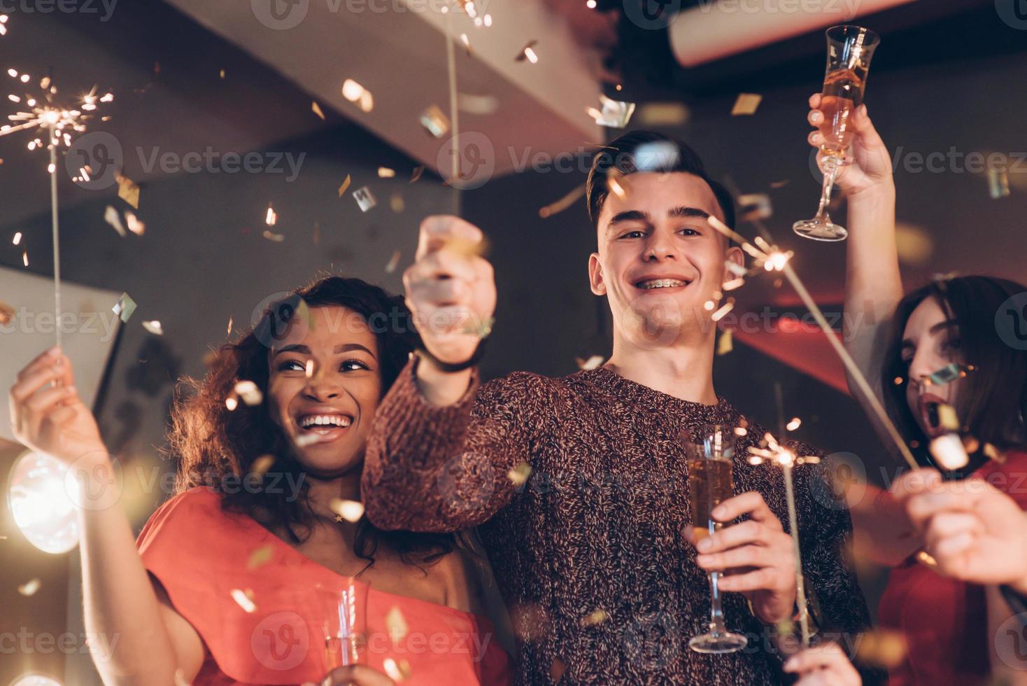 gioioso e felice. amici multirazziali festeggiano il nuovo anno e tengono in mano luci e bicchieri del Bengala con un drink foto