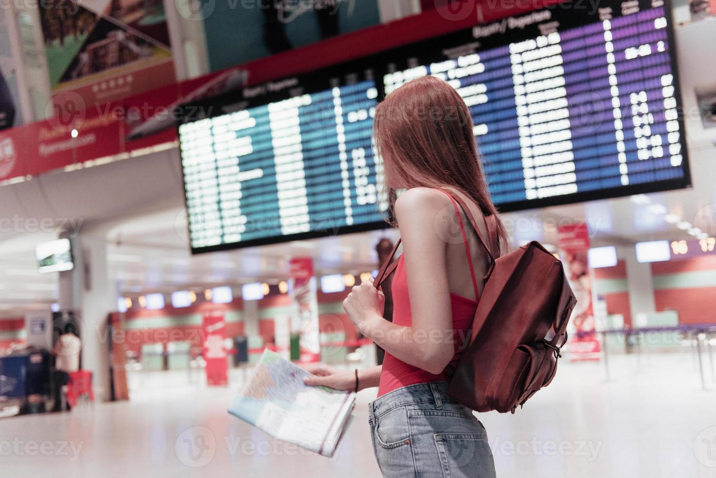 la giovane donna in aeroporto con la mappa in mano e la bacheca informativa sullo sfondo sta guardando di lato foto