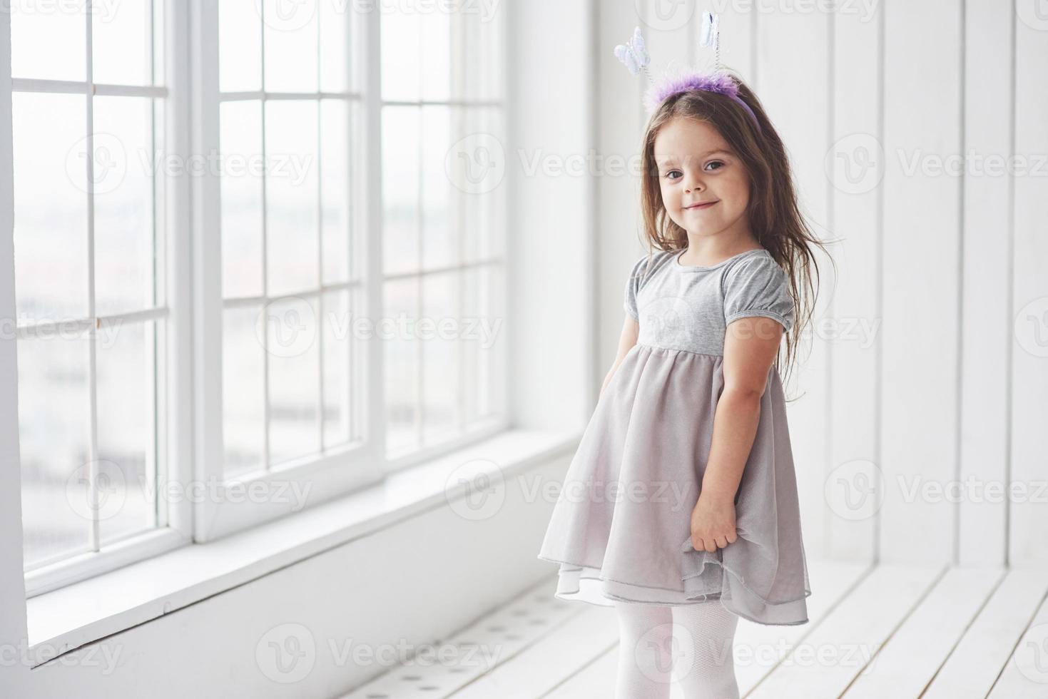 bambina carina in abito in piedi nella stanza bianca vicino alle finestre foto