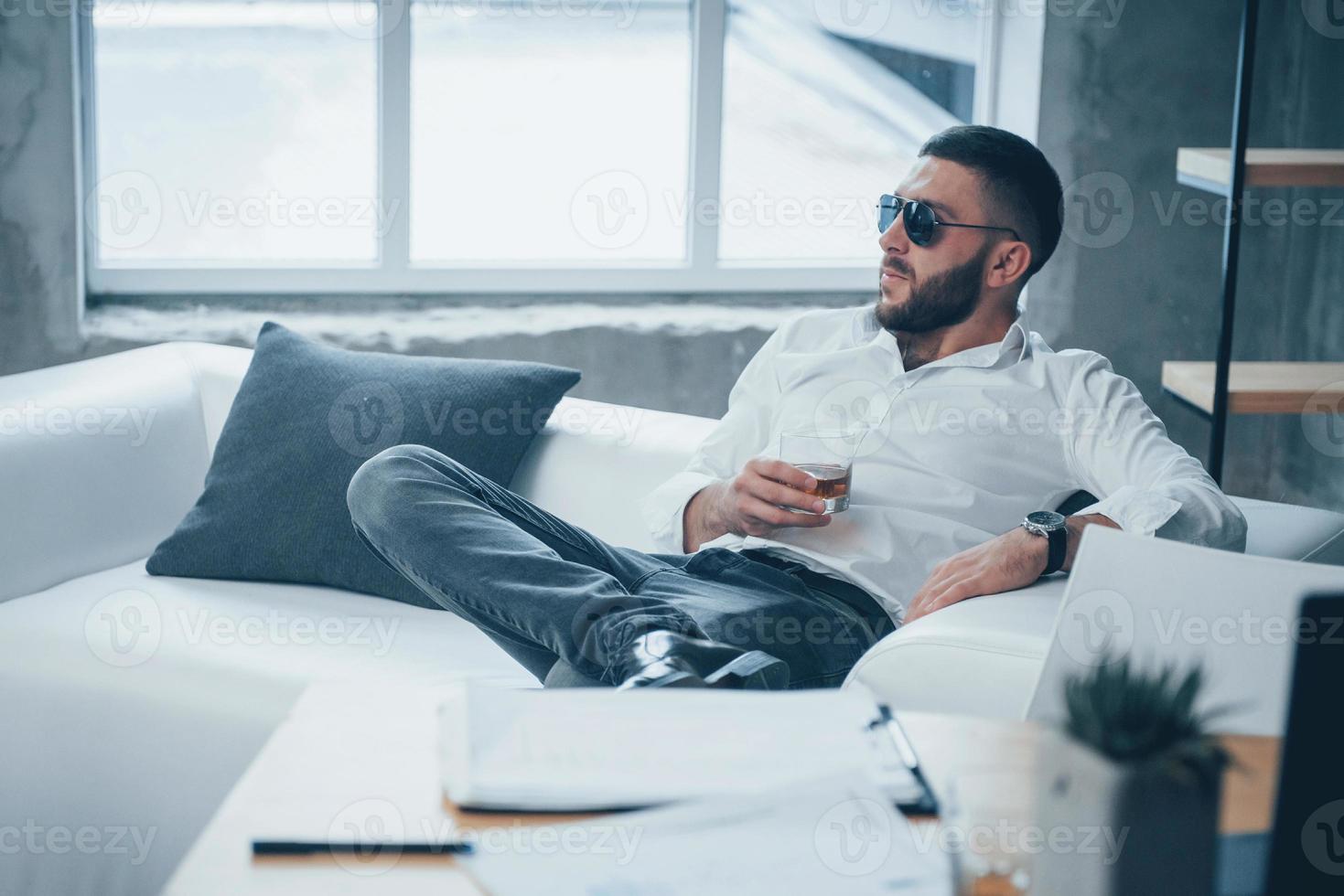 posa elegante. giovane uomo dai capelli corti in occhiali da sole seduto sul divano in ufficio foto