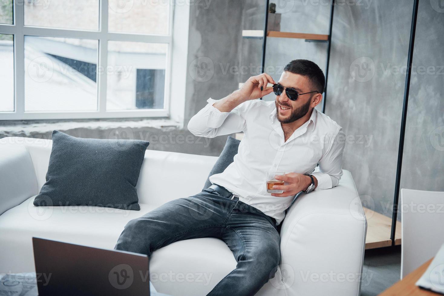 toccando gli occhiali e sorridendo. giovane uomo dai capelli corti in occhiali da sole seduto sul divano in ufficio foto