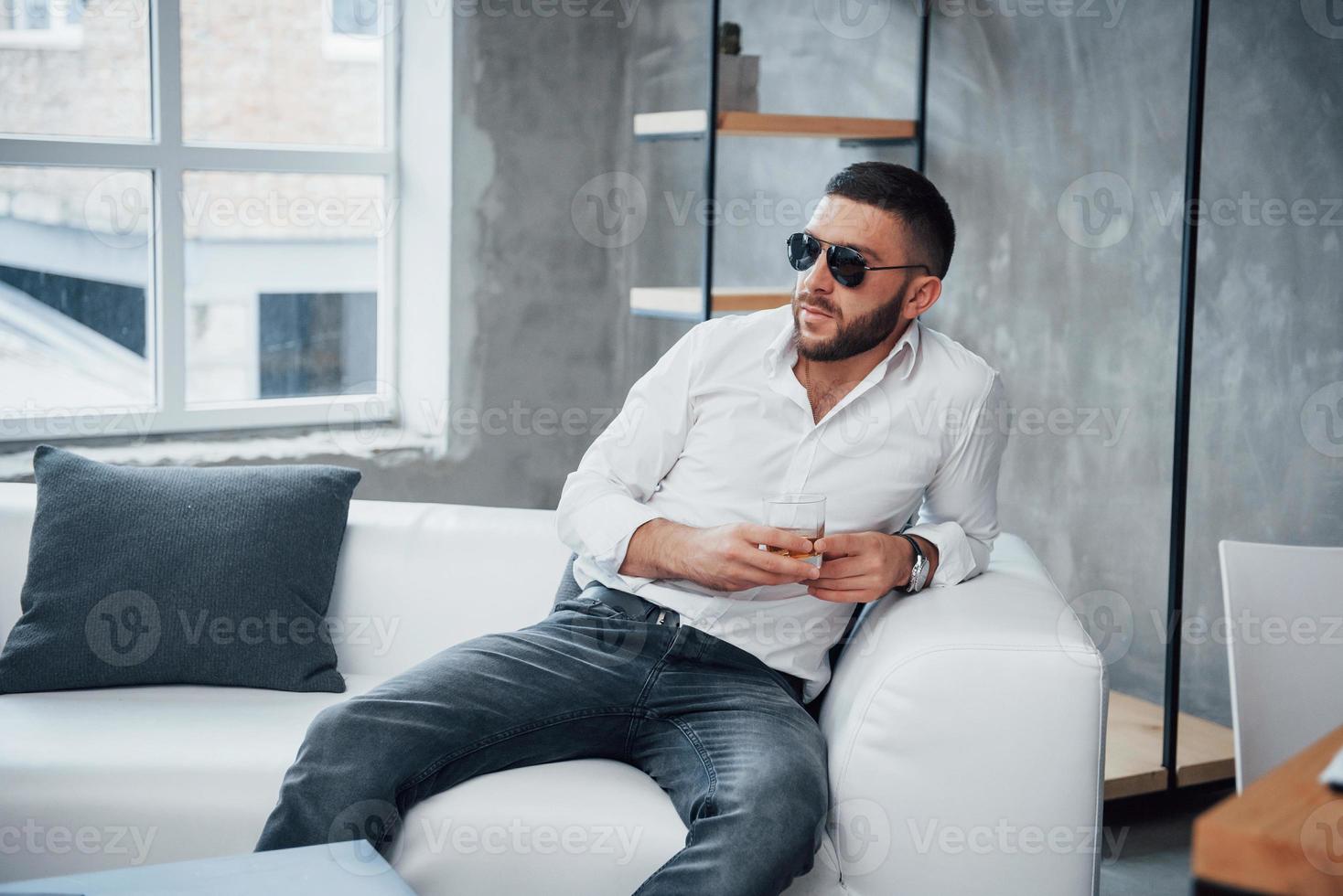 pensieri sul futuro. giovane uomo dai capelli corti in occhiali da sole seduto sul divano in ufficio foto