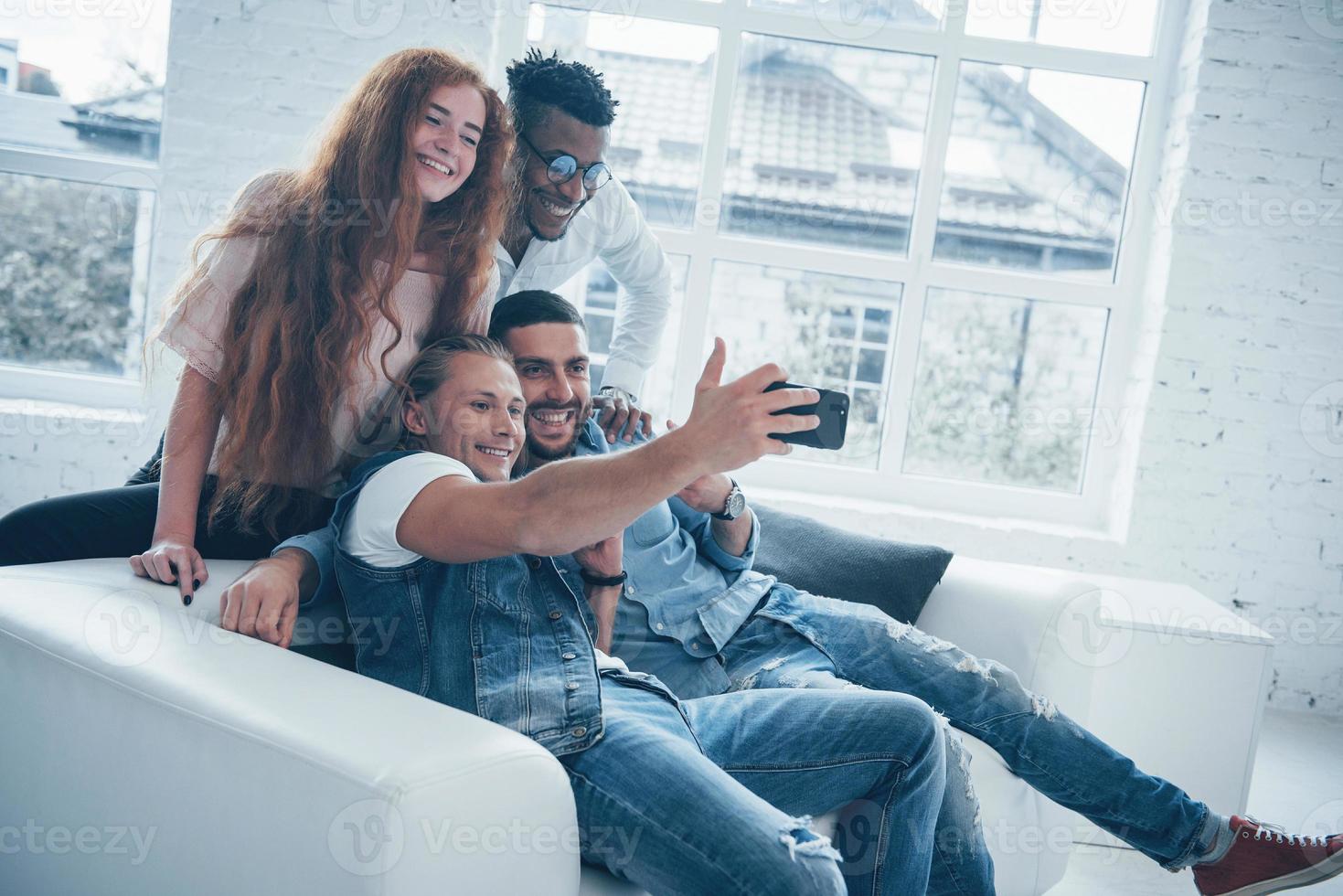 gli impiegati hanno una pausa. giovani amici allegri che si fanno selfie sul divano e interni bianchi foto