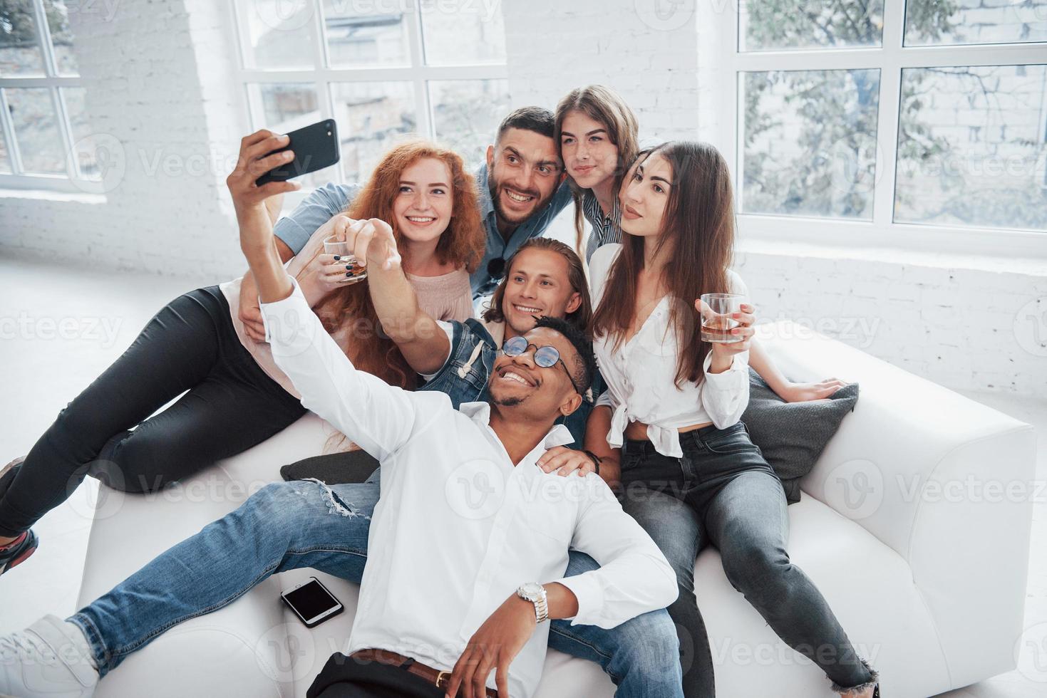 fare selfie. giovani amici allegri che si divertono e bevono nell'interno bianco foto