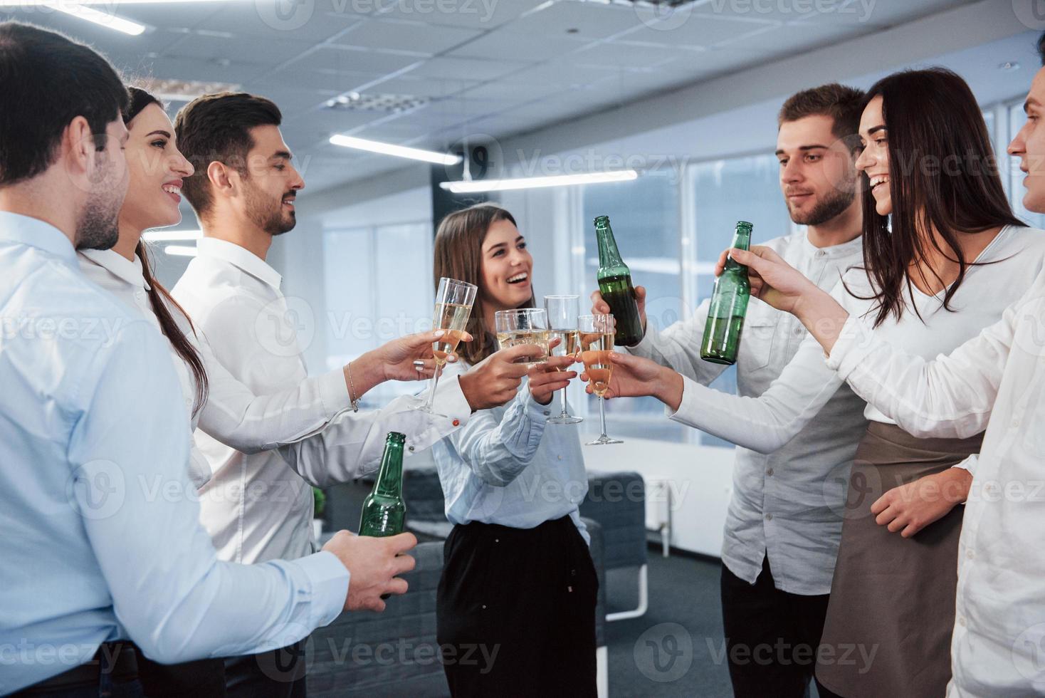 sorrisi sinceri. in piedi e bussando alle bottiglie e al bicchiere. nell'ufficio. i giovani festeggiano il loro successo foto