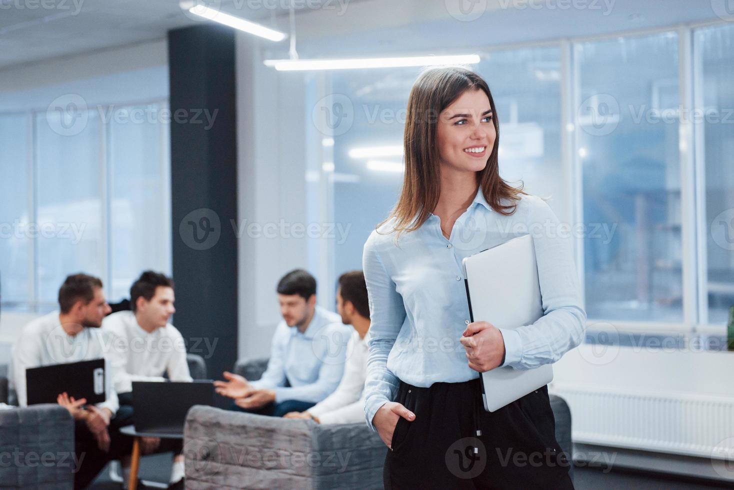 bruna è innamorata del suo lavoro. ritratto di giovane ragazza in piedi in ufficio con i dipendenti in background foto
