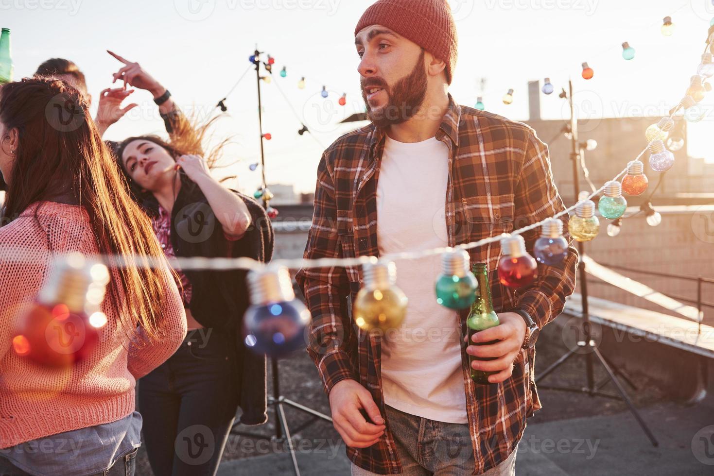 festa sul tetto. il giovane barbuto con il cappello rosso tiene la birra mentre i suoi amici si divertono foto