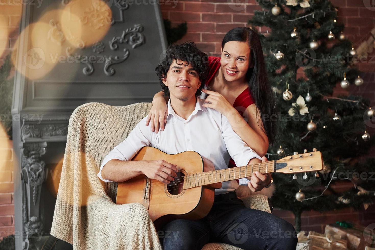 ritratto di bella coppia in vacanza. ragazzo attraente dai capelli ricci seduto sulla sedia con la chitarra acustica con l'albero di natale dietro. la ragazza in abito bianco sta abbracciando il suo ragazzo foto