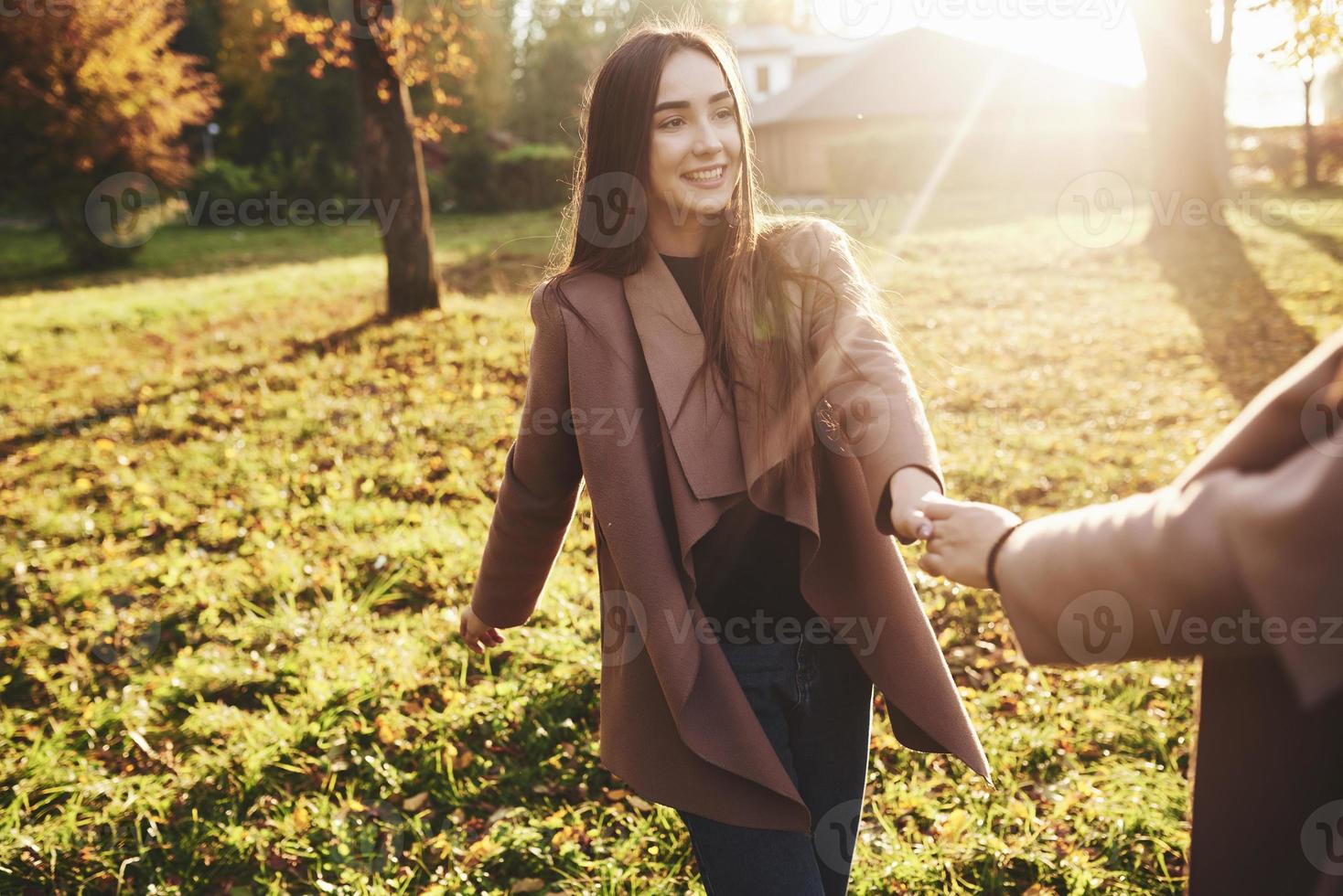 giovane, sorridente, bella ragazza bruna tiene la mano di qualcuno mentre cammina nel parco soleggiato autunnale su sfondo sfocato foto
