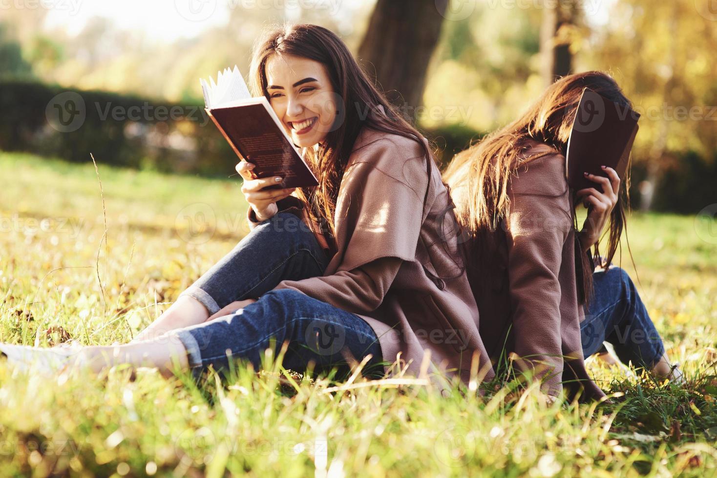 ridendo ragazza gemella bruna seduta di schiena sull'erba con sua sorella, che sta coprendo il viso con un libro marrone, indossando un cappotto casual nel parco soleggiato autunnale su sfondo sfocato foto