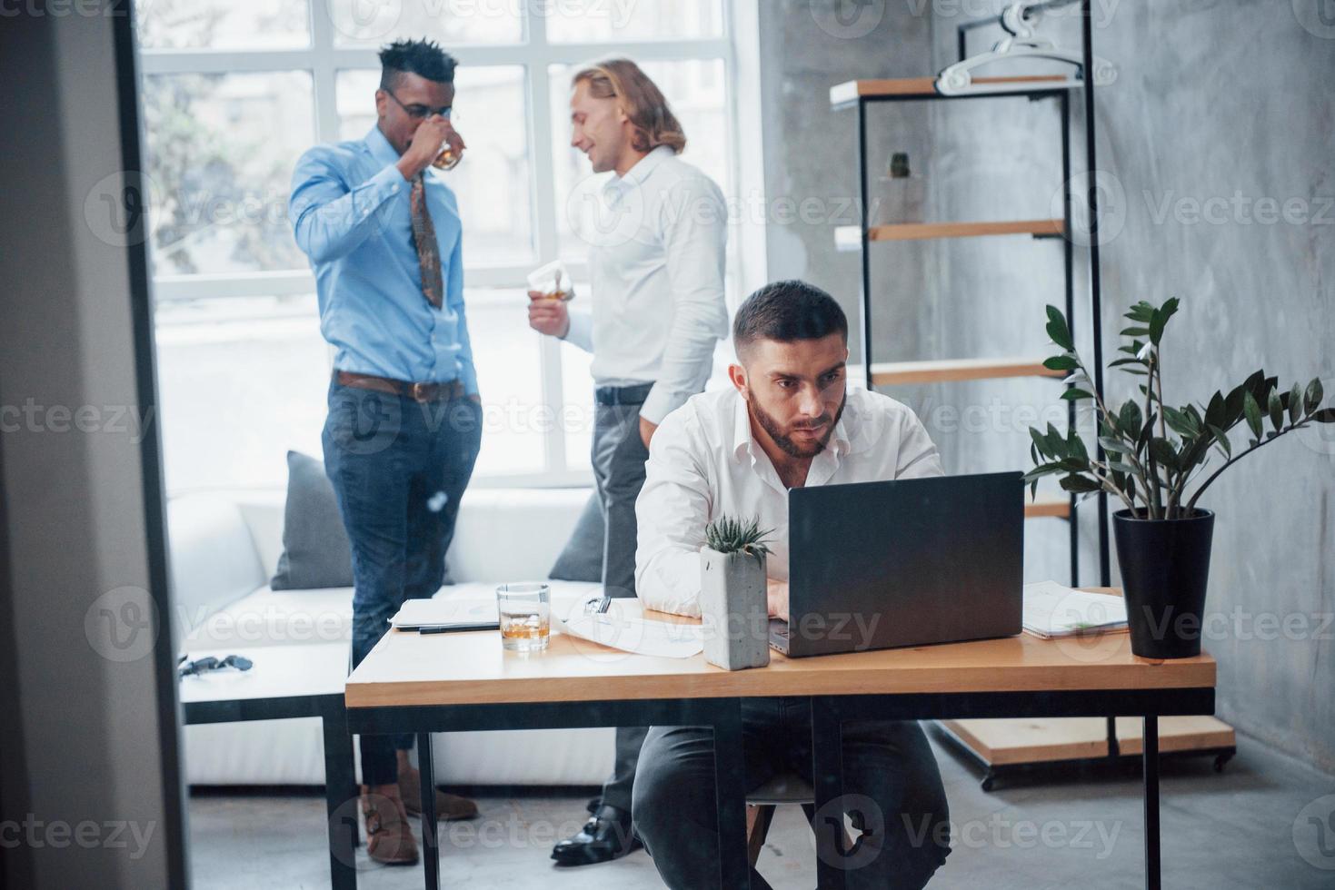 le persone hanno discussioni in background. il giovane uomo d'affari si siede sulla sedia davanti al computer portatile e lavora con i rapporti foto