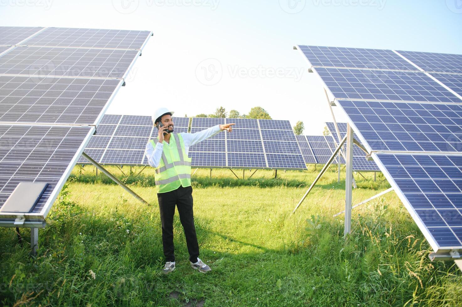 ritratto di giovane indiano uomo tecnico indossare bianca difficile cappello in piedi vicino solare pannelli contro blu cielo.industrial lavoratore solare sistema installazione, rinnovabile verde energia generazione concetto. foto