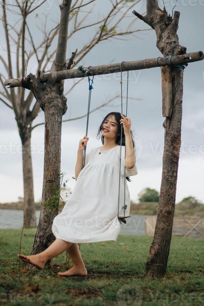 un' infanzia sognare di volante alto su un' swing nel un' parco. un' giovane ragazza nel un' bianca vestito cavalcate su un' swing foto