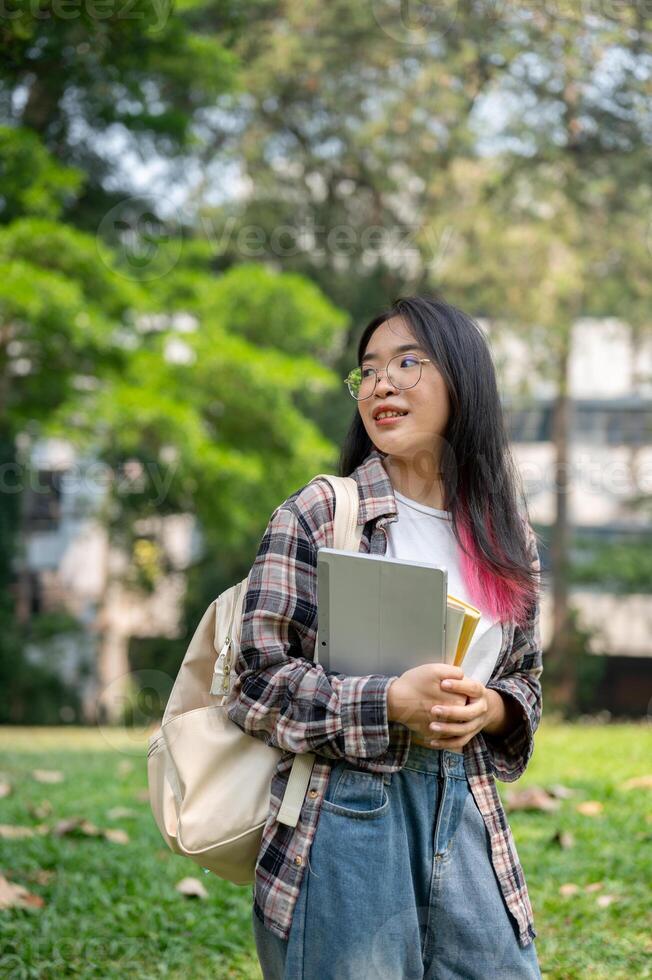 un' giovane asiatico femmina Università alunno trasporto un' zaino e roba, in piedi nel il città universitaria parco. foto