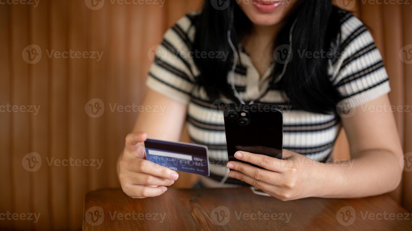 un' ritagliata Immagine di un asiatico donna Tenere un' smartphone e un' credito carta a un' tavolo nel un' caffè negozio foto