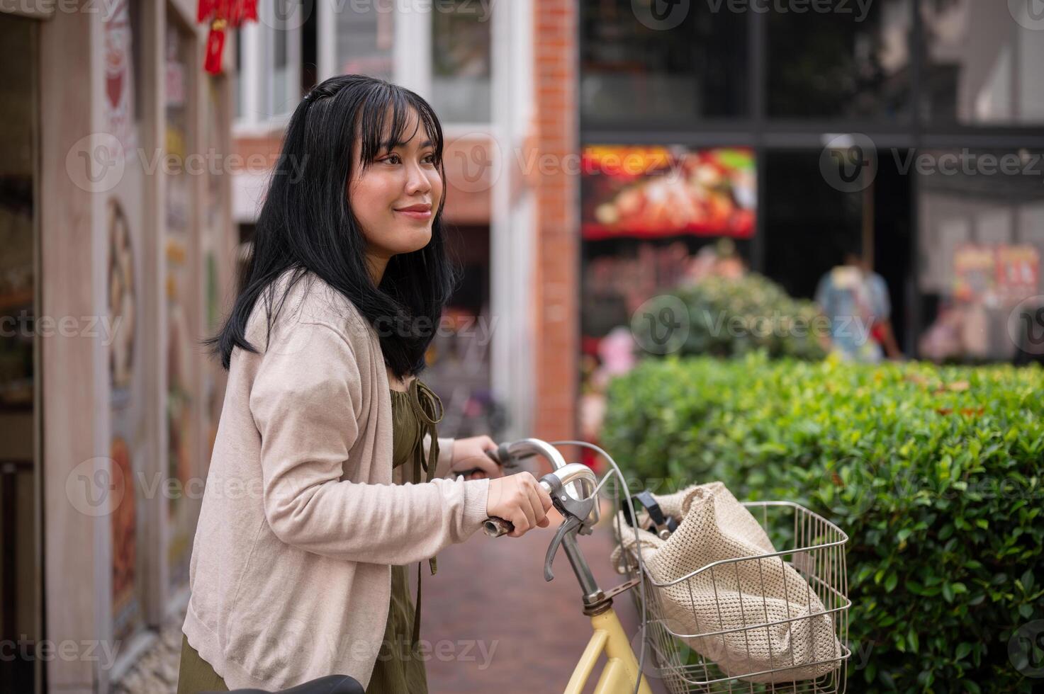 un' Bellissima, positivo asiatico donna nel un' carino vestito è a piedi e spingendo sua bicicletta nel il città. foto