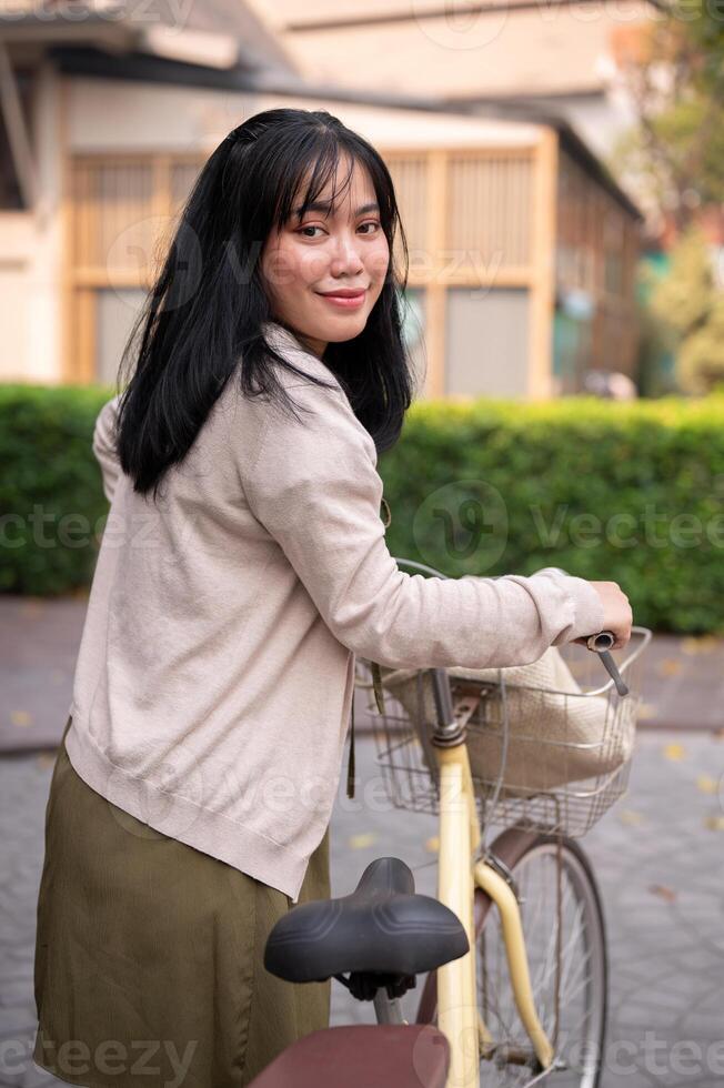 un' sorridente, contento giovane asiatico donna a piedi e spingendo sua bicicletta nel il città. foto