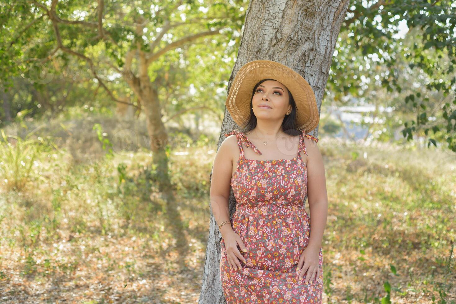 donna indossare cappello e vestito con fiori ammirazione natura nel un' pubblico parco vicino per un' albero. soleggiato estate giorno. foto