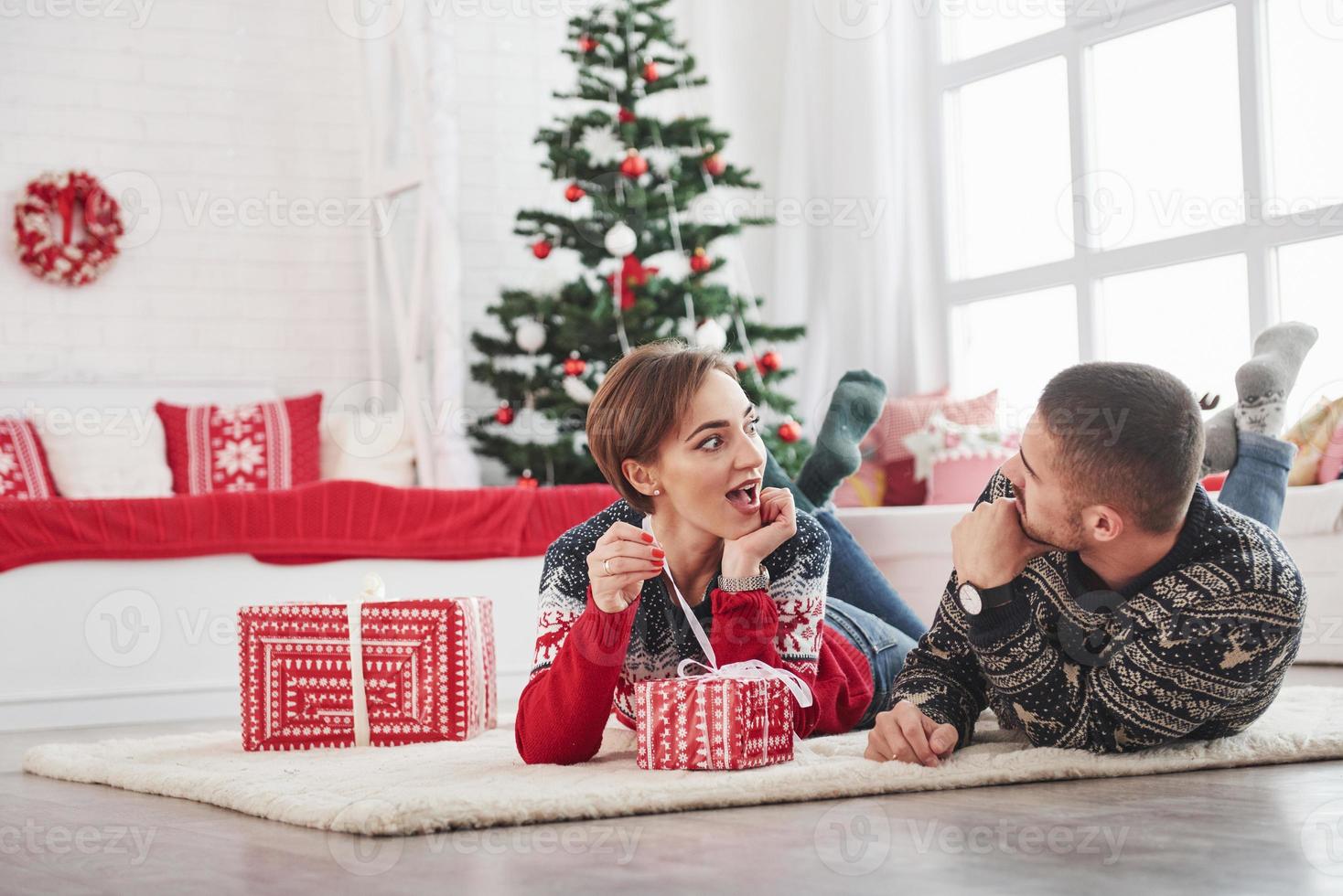 la ragazza è entusiasta del tipo di regalo all'interno della scatola. adorabile giovane coppia sdraiata sul soggiorno con albero di vacanza verde sullo sfondo foto