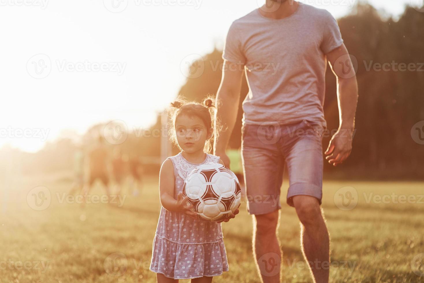 chiedendosi fino a che punto questa ragazza può calciare quel pallone. foto di papà con sua figlia in un bellissimo prato e boschi sullo sfondo