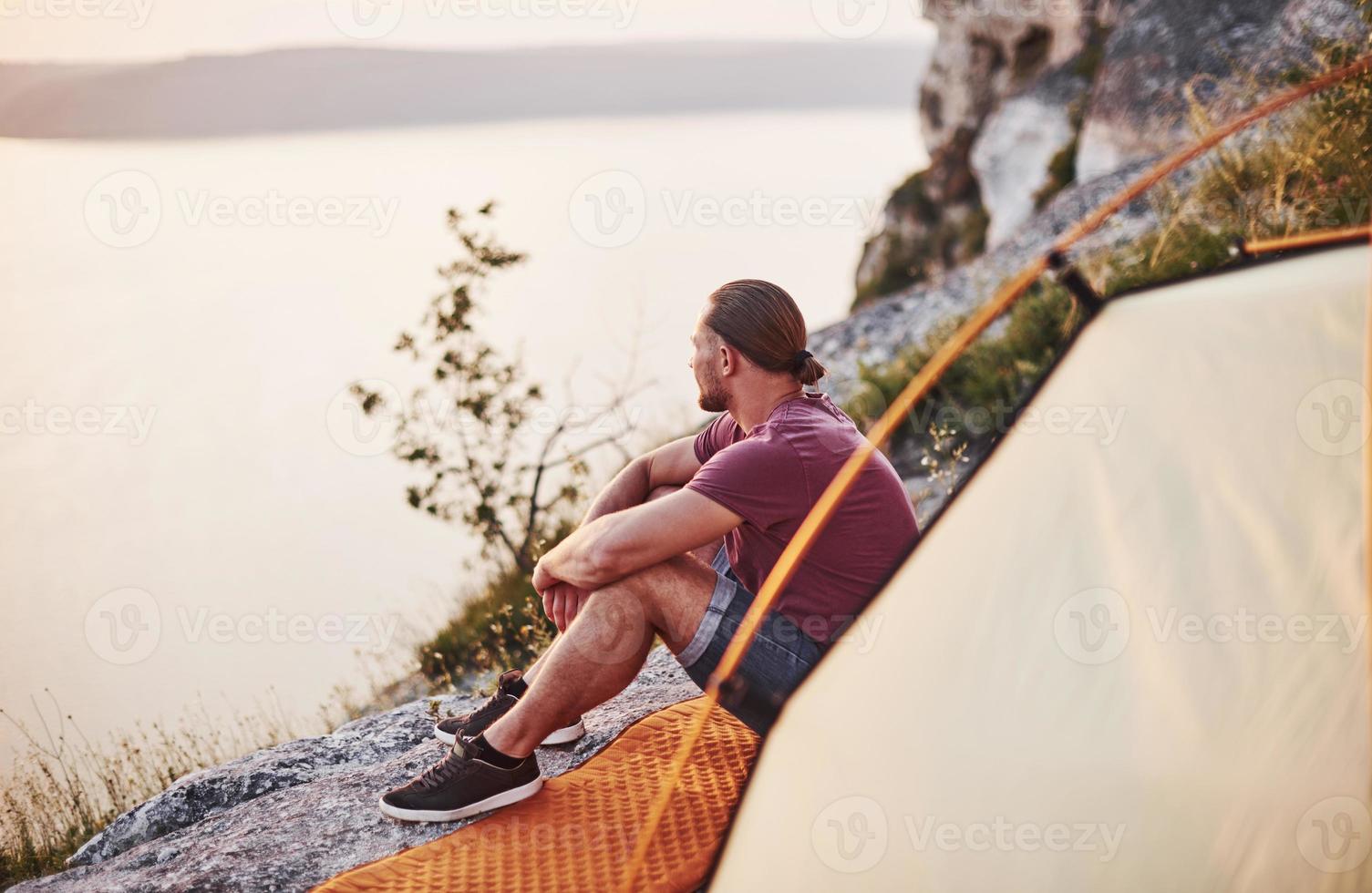 turista in pantaloncini e maglietta seduto sul bordo della montagna nella parte anteriore della tenda foto