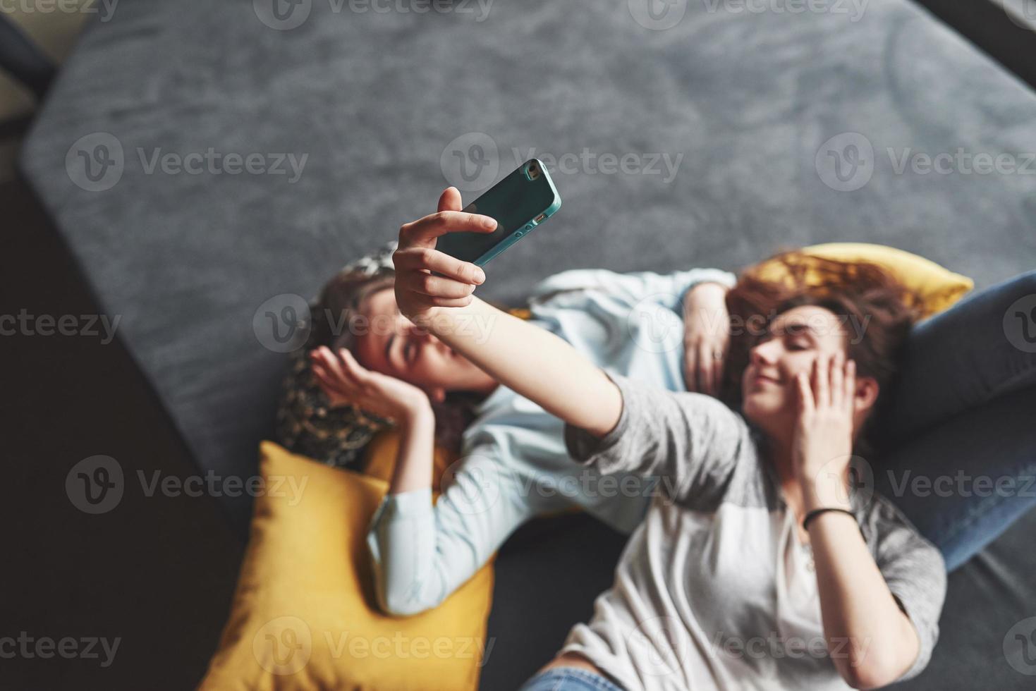 due sorelle gemelle sorridenti carine che tengono smartphone e fanno selfie. le ragazze si sdraiano sul divano in posa e gioia foto