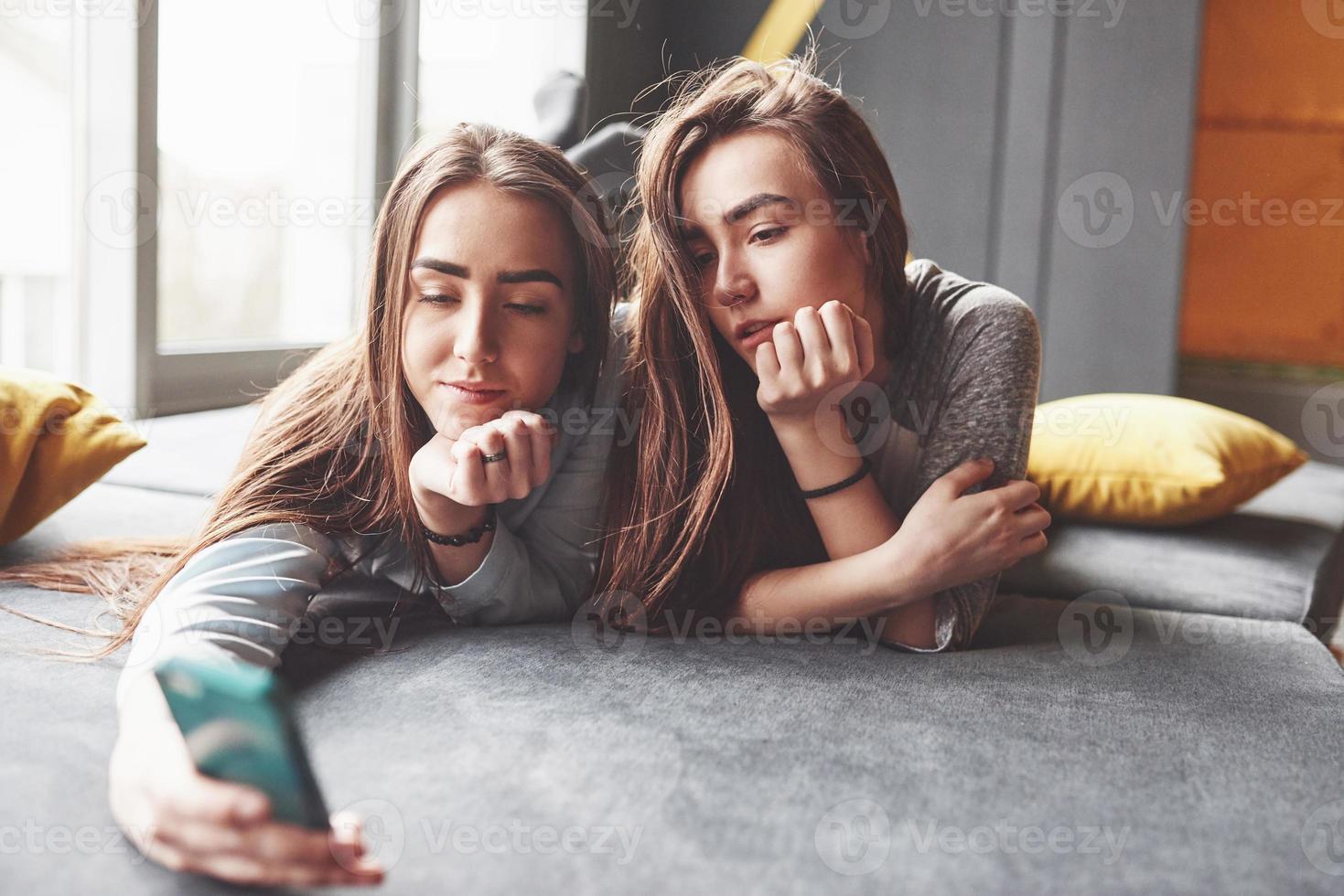 due sorelle gemelle sorridenti carine che tengono smartphone e fanno selfie. le ragazze si sdraiano sul divano in posa e gioia foto