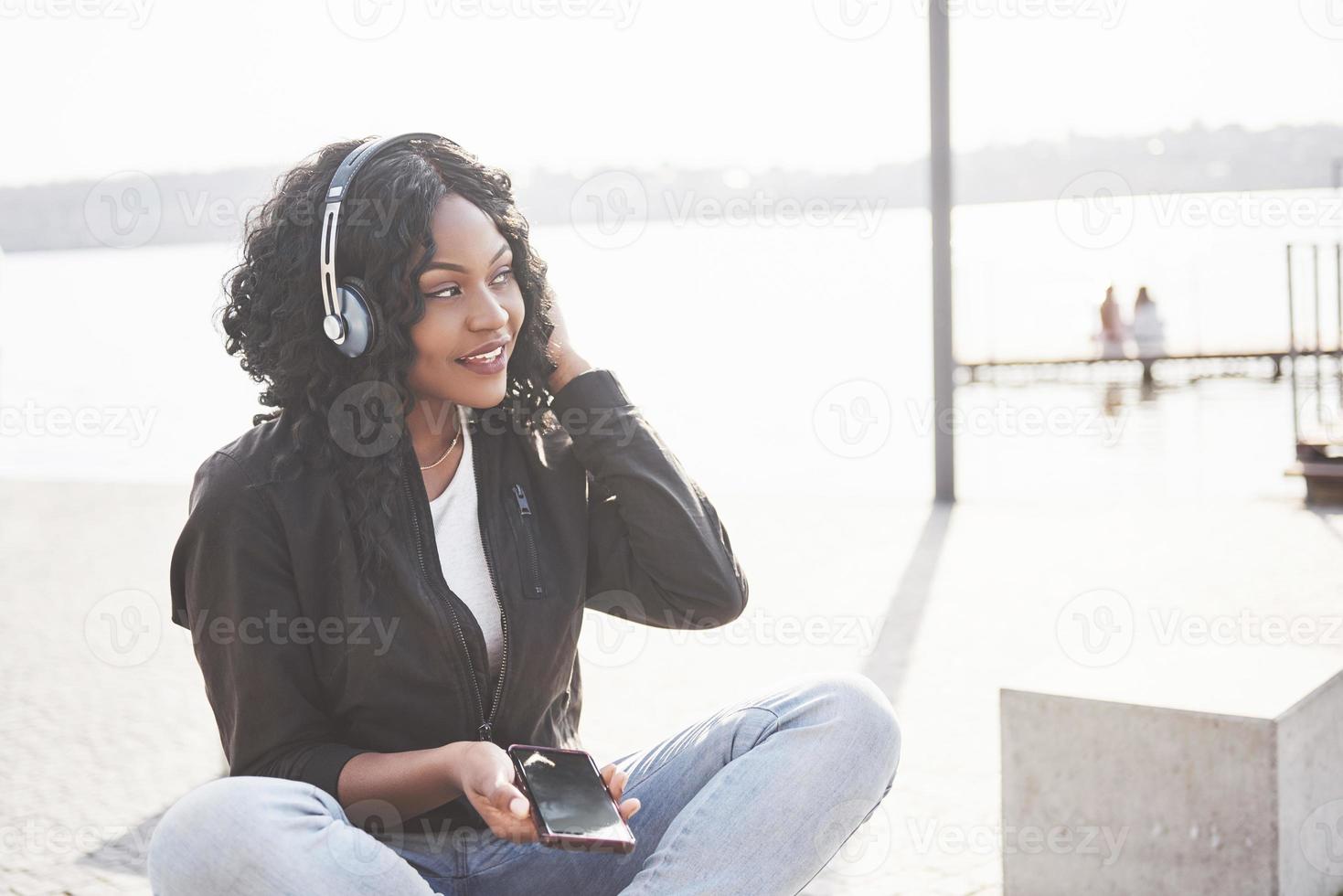 ritratto di una giovane e bella ragazza afroamericana seduta sulla spiaggia o sul lago e ascoltando musica nelle sue cuffie foto