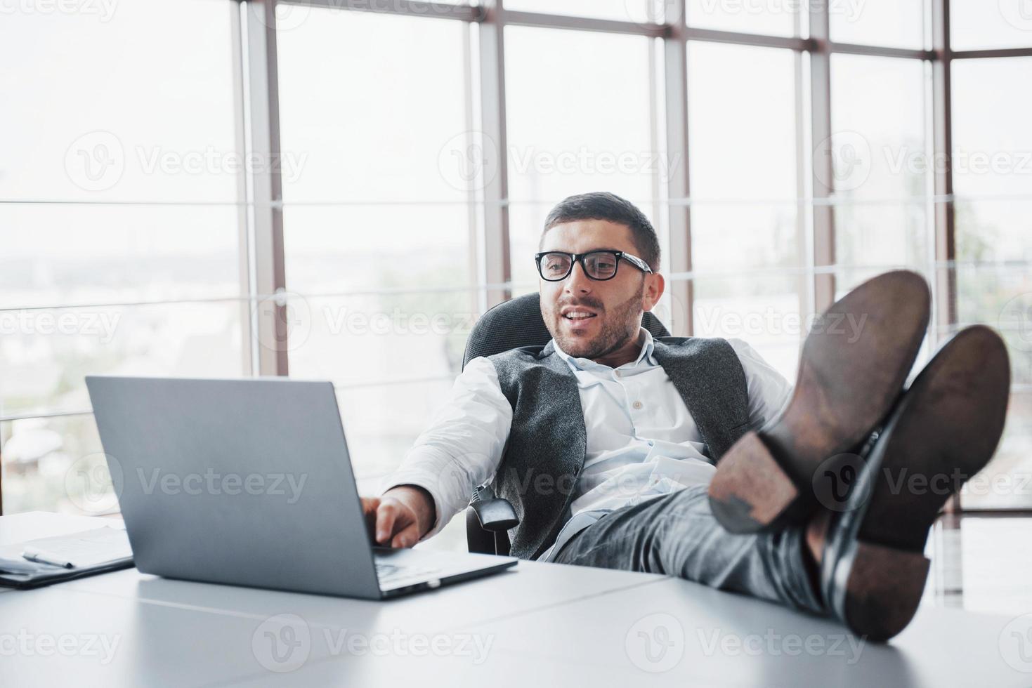 bellissimo giovane uomo d'affari con gli occhiali che tiene le gambe sul tavolo guardando un laptop in ufficio foto