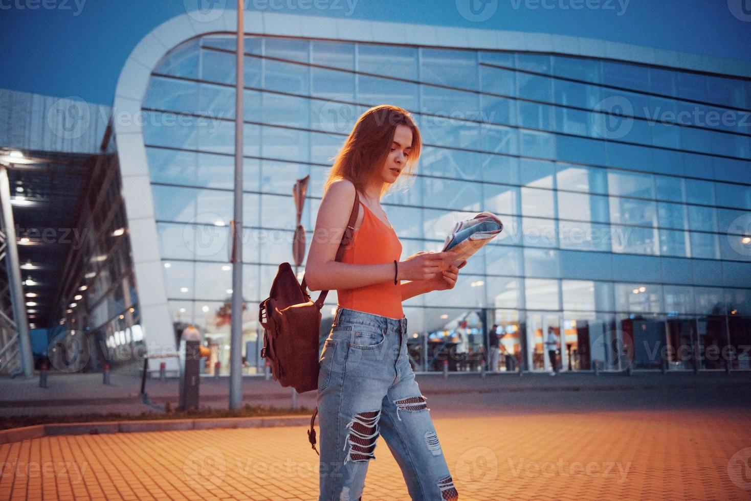 la ragazza costa di notte vicino al terminal dell'aeroporto o della stazione e legge la mappa della città e cerca un hotel. simpatico turista con zaini determina il concetto di viaggio foto