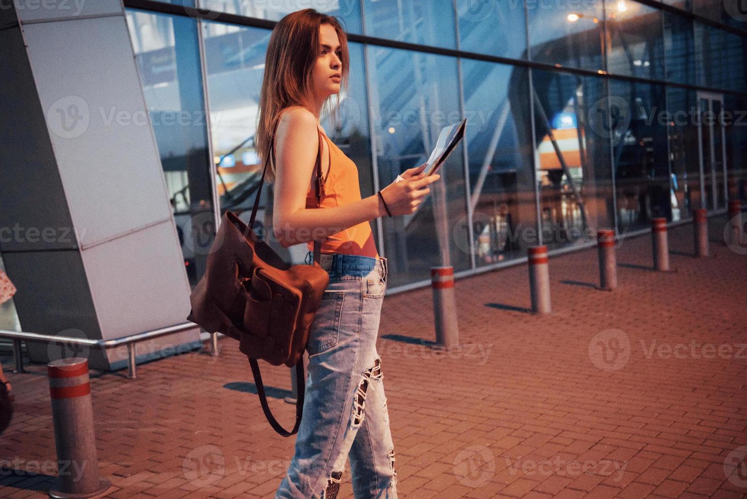 una giovane bella ragazza con uno zaino dietro le spalle in piedi sulla strada vicino a un aeroporto. è appena arrivata dal resto e molto felice foto
