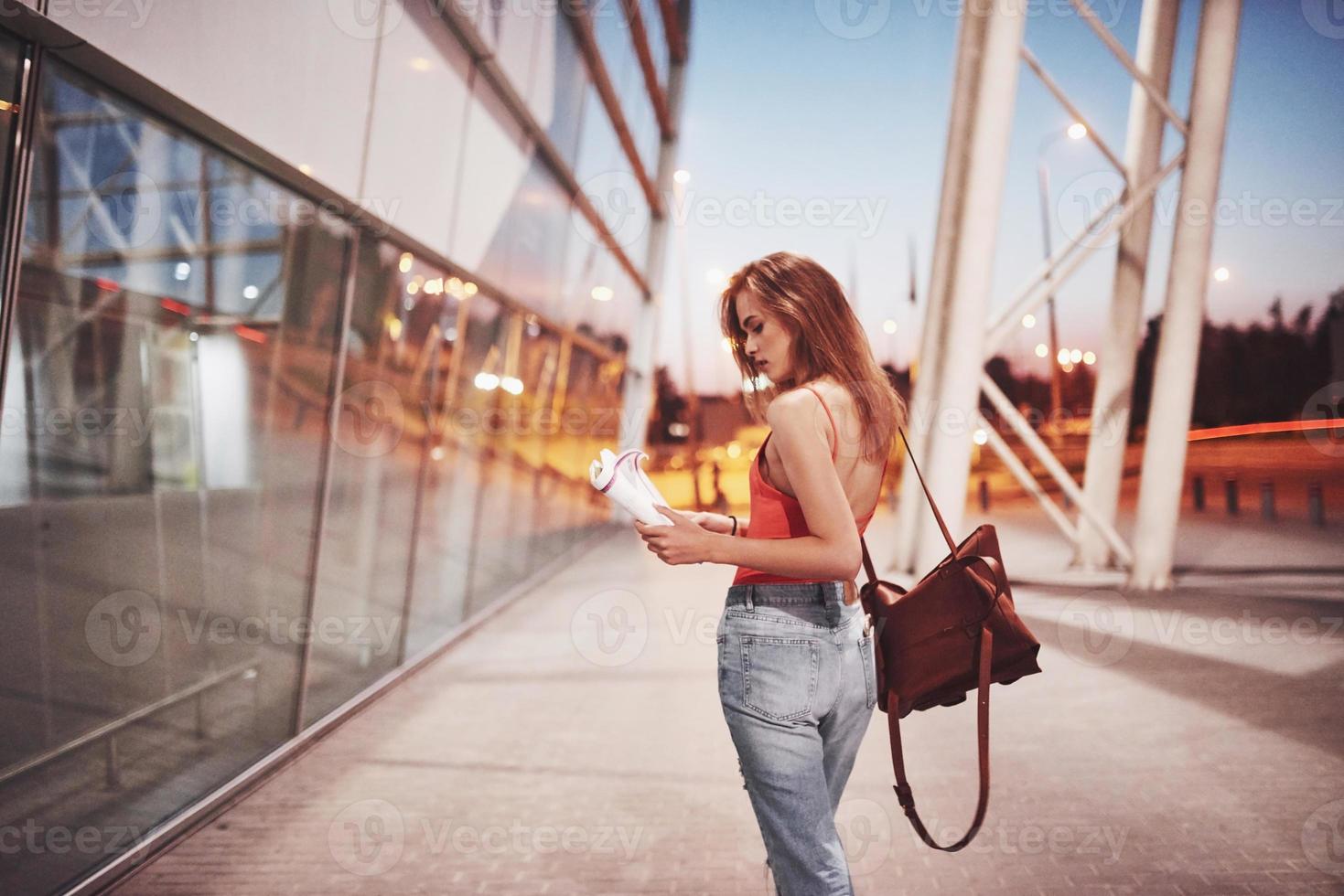 una giovane bella ragazza con uno zaino dietro le spalle in piedi sulla strada vicino a un aeroporto. è appena arrivata dal resto e molto felice foto