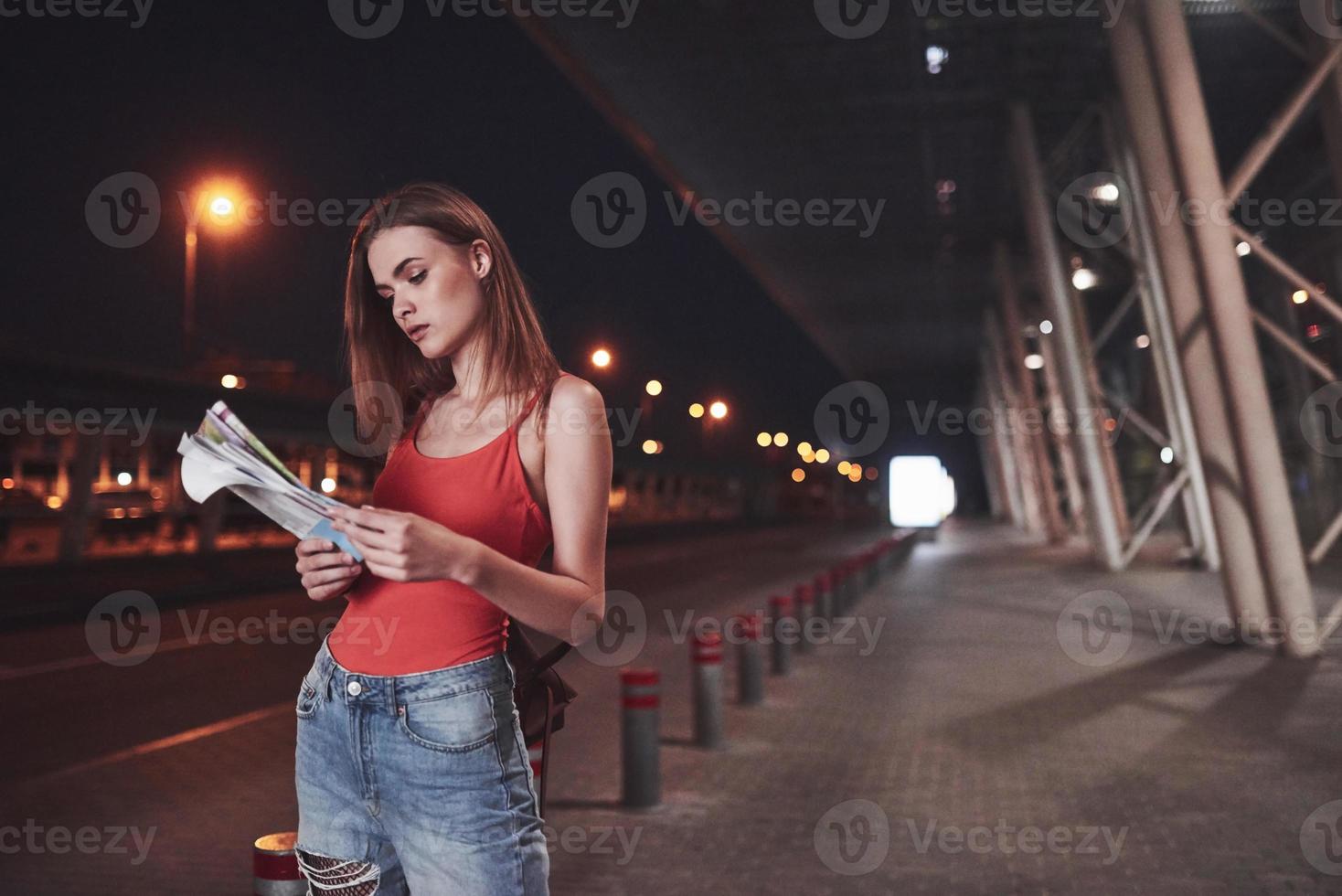 la ragazza costa di notte vicino al terminal dell'aeroporto o della stazione e legge la mappa della città e cerca un hotel. simpatico turista con zaini determina il concetto di viaggio foto