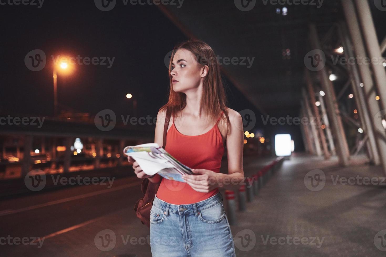 la ragazza costa di notte vicino al terminal dell'aeroporto o della stazione e legge la mappa della città e cerca un hotel. simpatico turista con zaini determina il concetto di viaggio foto