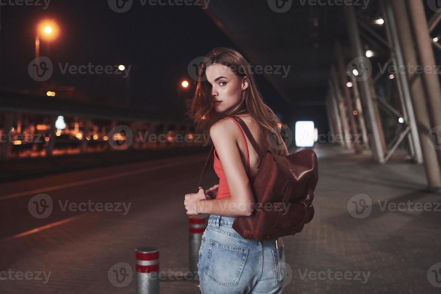 una giovane bella ragazza con uno zaino dietro le spalle si trova per strada vicino a un aeroporto o una stazione ferroviaria in una calda serata estiva. è appena arrivata e aspetta un taxi o i suoi amici foto