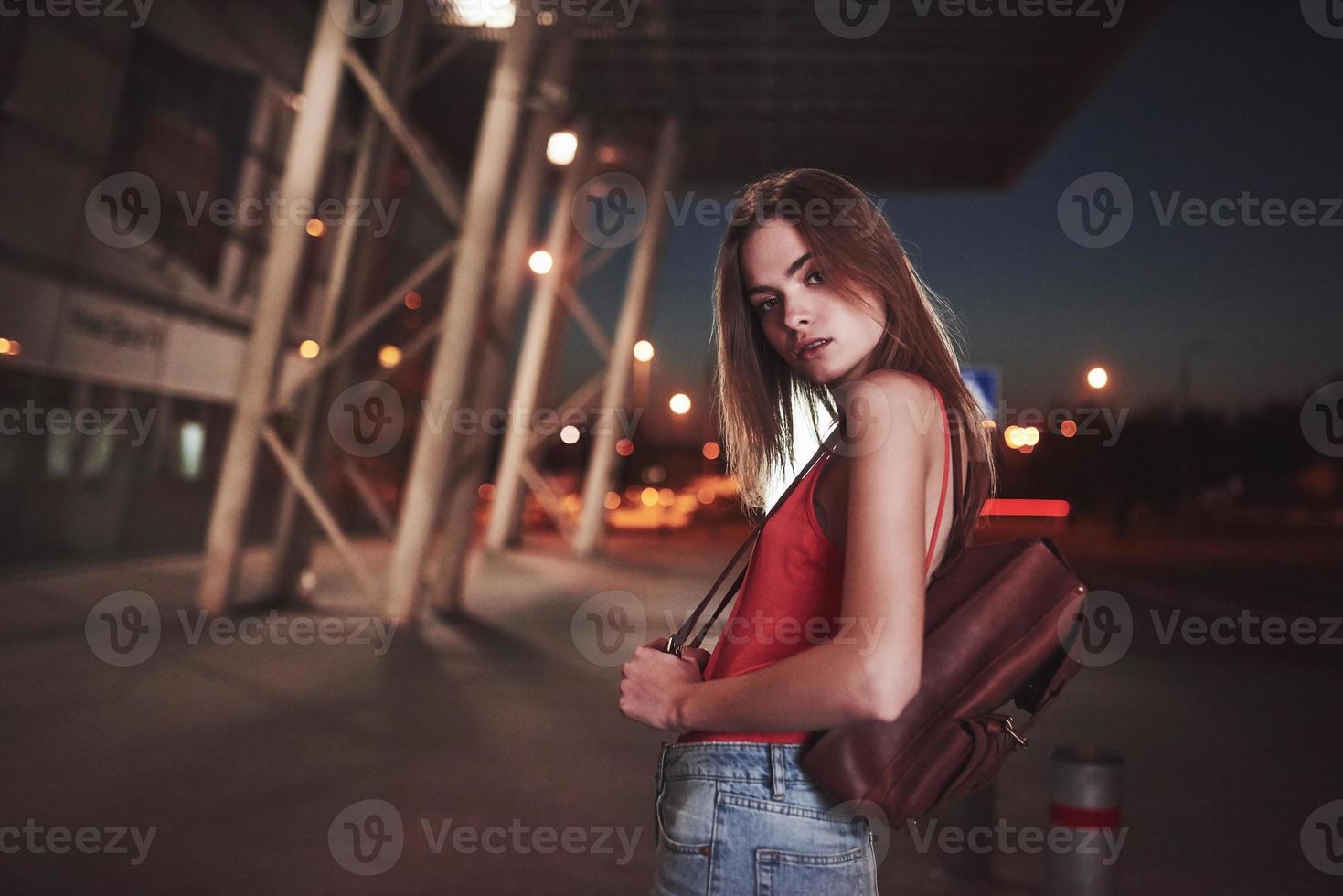 una giovane bella ragazza con uno zaino dietro le spalle si trova per strada vicino a un aeroporto o una stazione ferroviaria in una calda serata estiva. è appena arrivata e aspetta un taxi o i suoi amici foto