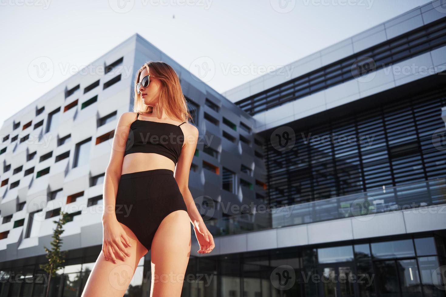 giovane donna sexy in occhiali da sole e costume da bagno nero è in piedi sullo sfondo degli edifici della città durante il tramonto foto