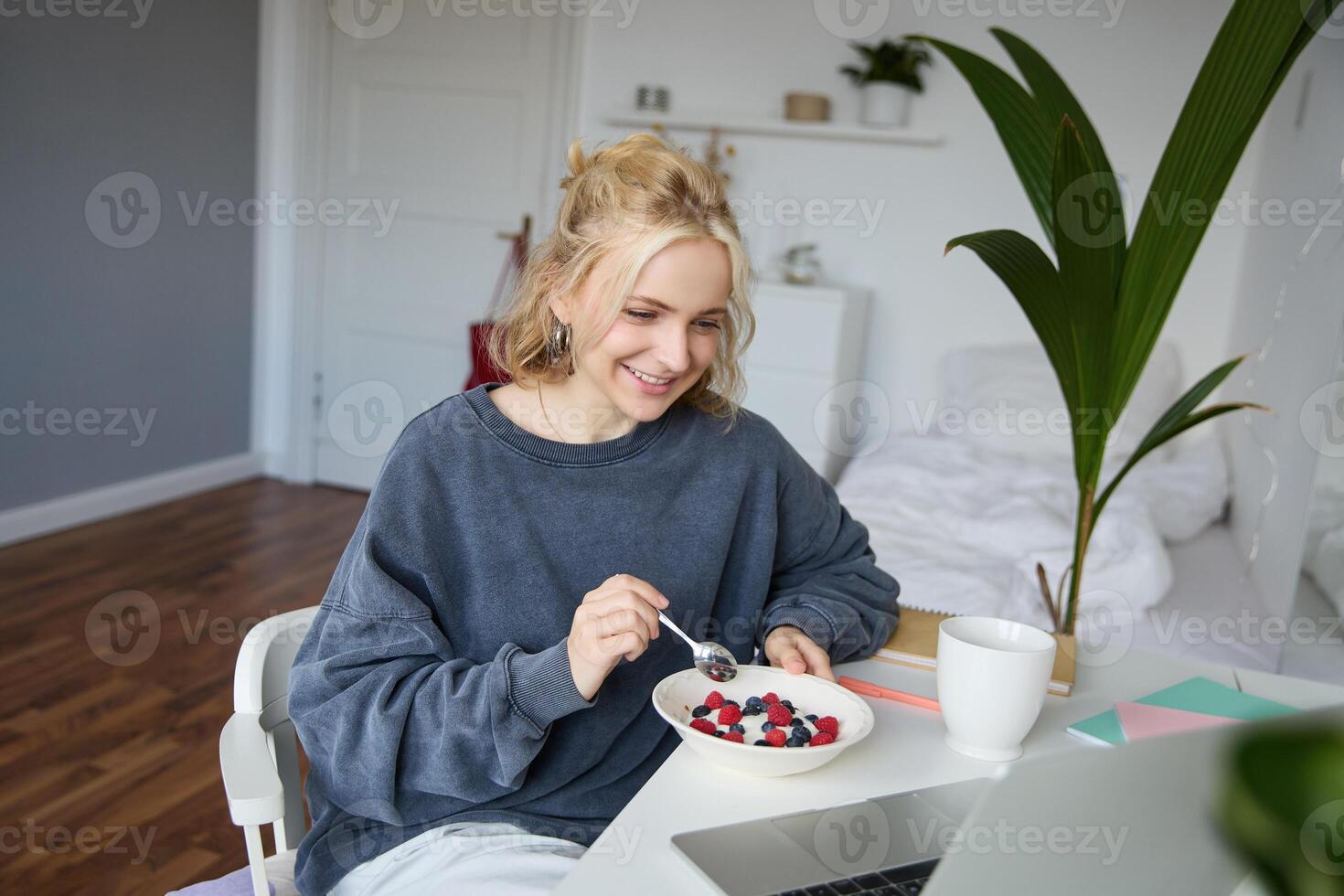 ritratto di sorridente biondo giovane donna, mangiare nel davanti di computer portatile, Guardando S in linea mentre avendo colazione, godendo dolce, seduta nel Camera da letto foto