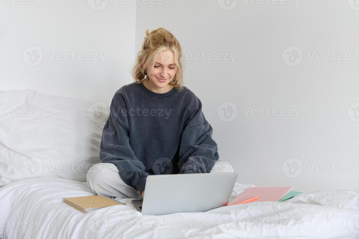 Immagine di contento giovane donna, alunno e-learning a partire dal casa, Collegare per in linea corso su sua computer portatile, si siede su letto con i Quaderni, sorridente e guardare contento foto