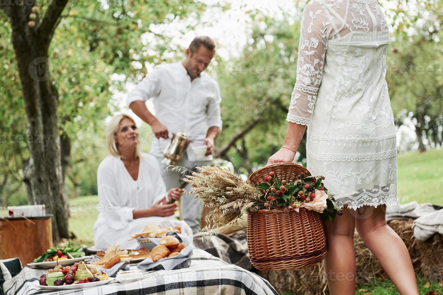 pronto per la cena. tre amici vicino al tavolo con cibo all'aperto. la ragazza tiene un cesto con piante secche foto