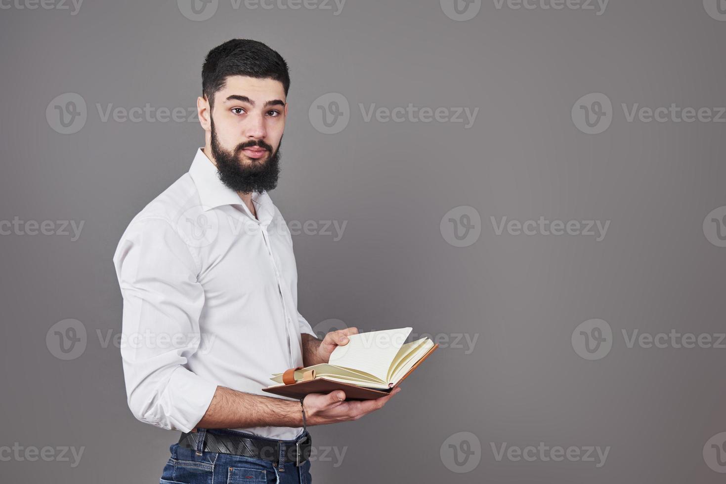 ritratto di un giovane barbuto che indossa una camicia bianca e tiene in mano un'agenda aperta e una penna. uno sfondo grigio muro foto