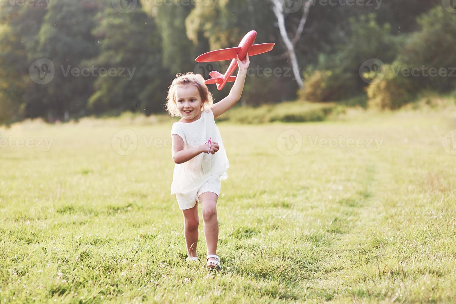 bambina felice che corre sul campo con un aereo giocattolo rosso nelle loro mani. alberi sullo sfondo foto