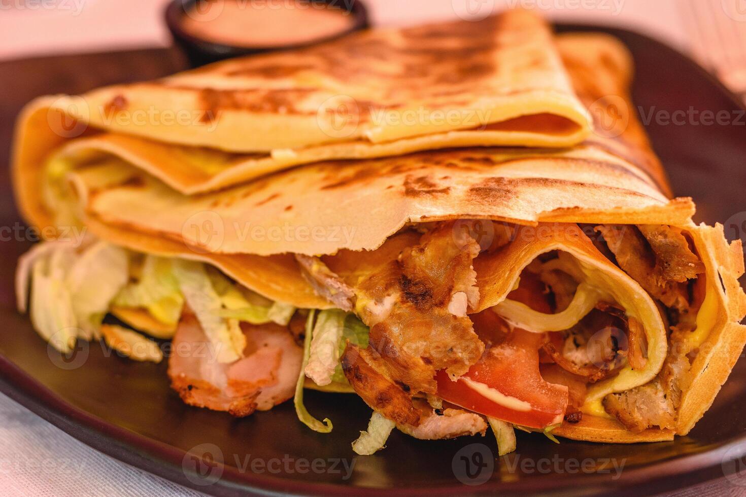 vicino su grande pancake pieno con pollo con formaggio, Bacon, pomodoro, lattuga, salsa su un' nero piatto nel greco taverna. magro Pancakes, crepes. selettivo morbido messa a fuoco foto