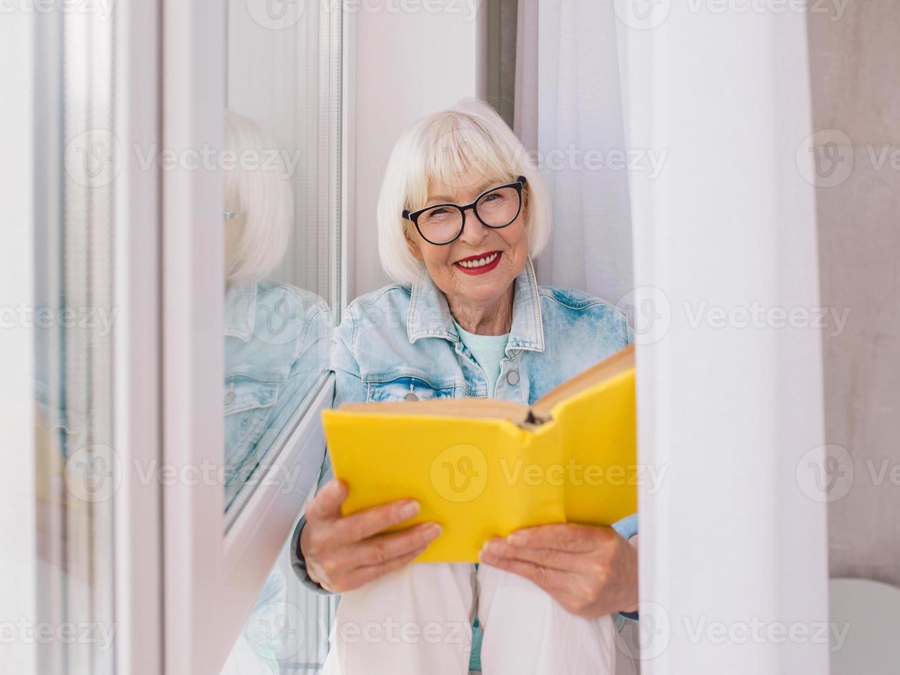 donna anziana con i capelli grigi che legge un libro vicino alla finestra a casa. educazione, pensione, anti età, concetto di lettura foto